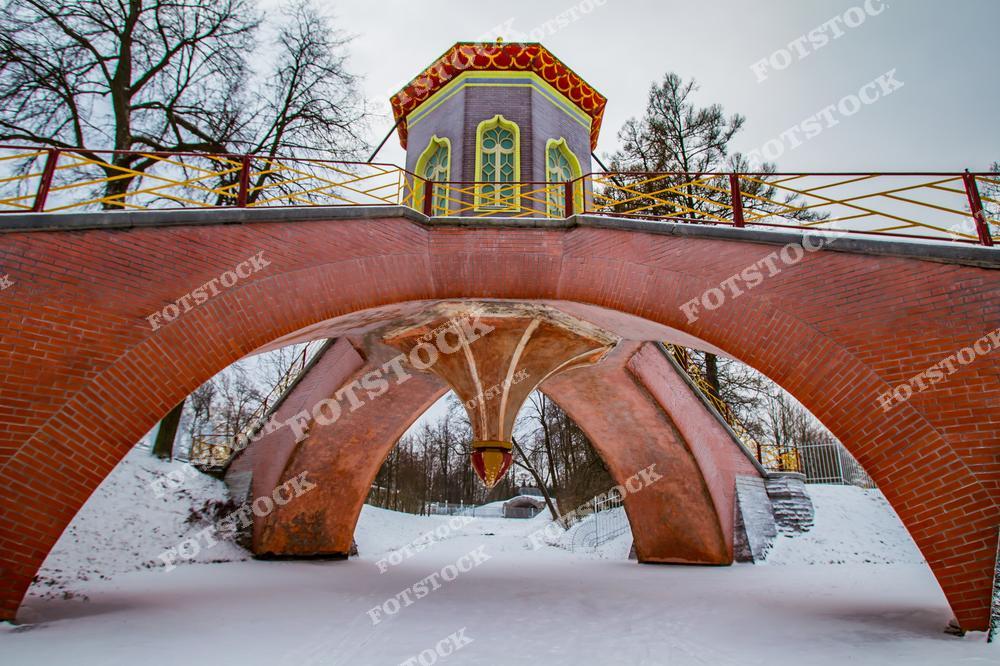 На фотографии запечатлен Крестовый мост, расположенный в Александровском парке Царского Села. Это яркий образец декоративной архитектуры, построенный из красного кирпича с арочными пролетами и небольшим павильоном наверху. Зимой мост выглядит особенно живописно, выделяясь на фоне снежного покрова и голых деревьев. Мост является важным элементом ландшафта парка и привлекает внимание своей уникальной конструкцией и исторической ценностью.